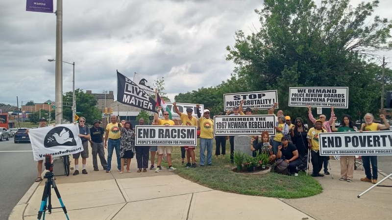 Activsts poose for a group shot, fists raised and holding People’s Organization for Progress flags with various slogans, as we as a Black Lives Matter flag and a Veterans for Oeace flag.