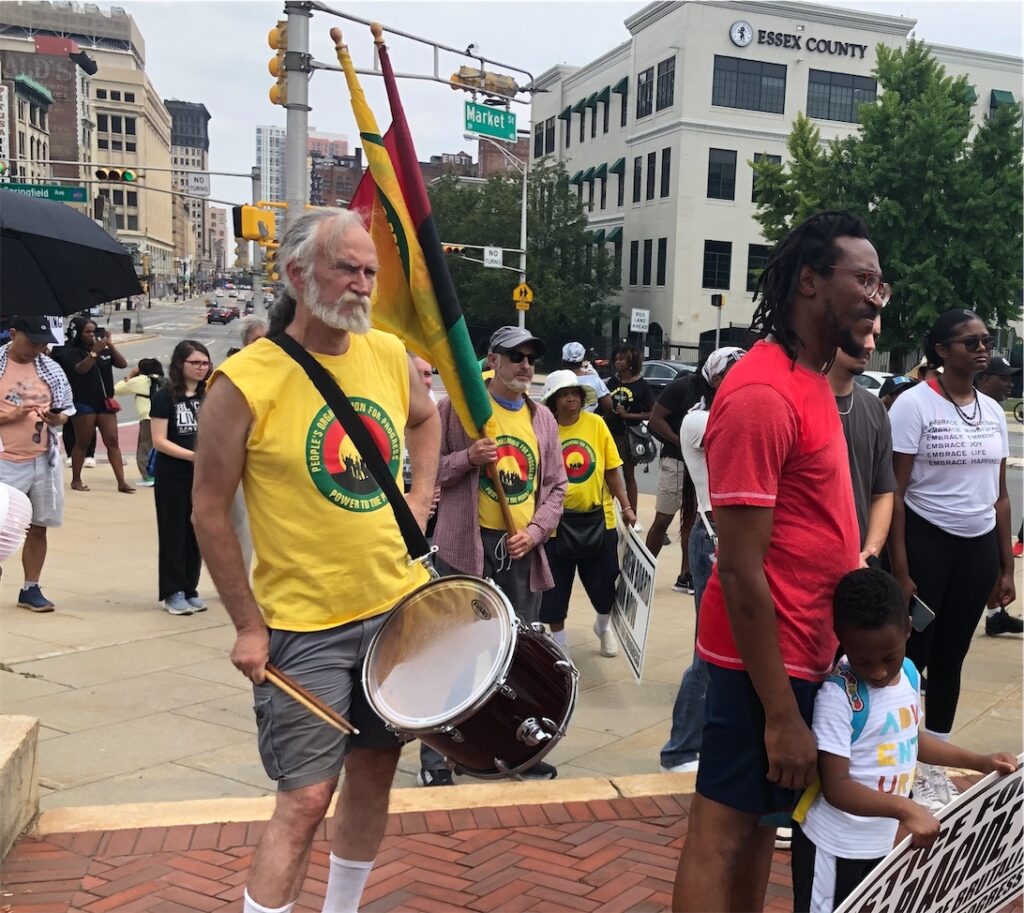 activists gather across the street from an Essex County building in Newark, N.J.; man in red shirt is with a boy holding a POP sign calling for justice, 3 people wearing POP T-shirts, 1 with a drum, 1 with 2 flags (POP and PanAfrican), and 1 with a sign