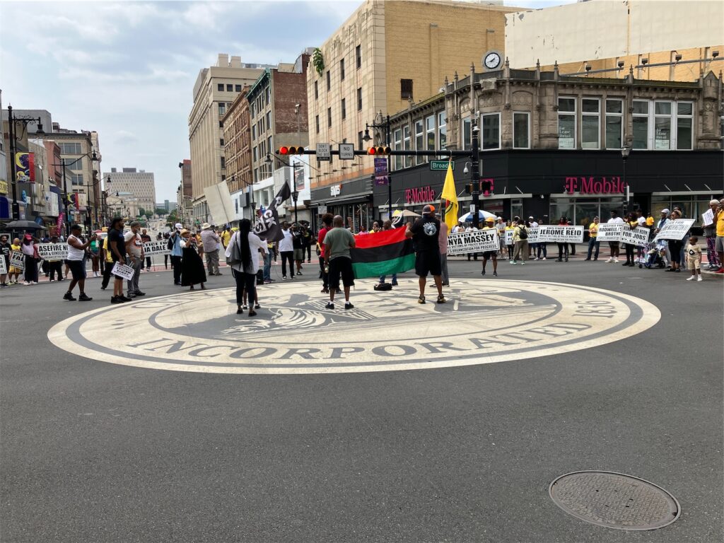 half of Broad and Market streets’ intersection, Newark, N.J., with PanAfrican, Black Lives Matter, and People’s Organization for Progress flags in the center, surrounded by activists, many holding POP signs with different text on each