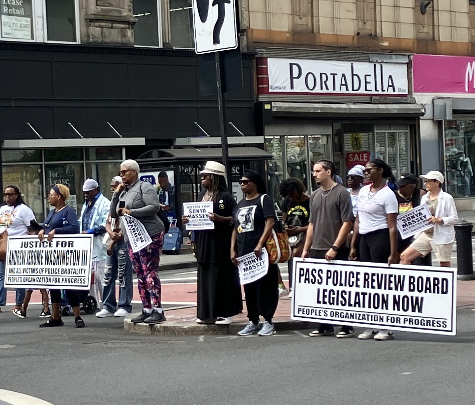 Activists at rally in Newark intersection, holding signs calling for Justice for Sonya Massey, passing Police Review Board legislation, and justice for Andrew Jerome Washington III.