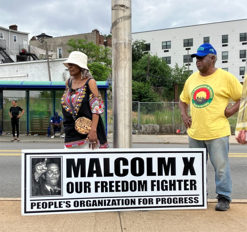Activists—woman in African print blouse and white bucket hat, and man in jeans, yellow POP T-shirt, and blue baseball cap, stand behind a People’s Organization for Progress sign with a photo of Malcom X that also says “MALCOM X: OUR FREEDOM FIGHTER”.