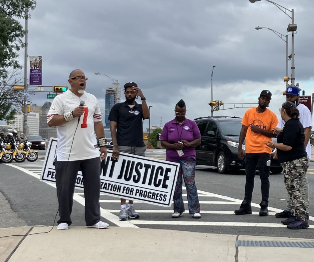 Black activists, one with a microphone, stand in a Newark crosswalk, just off the curb. Visible on one People ’s Organization for Progress sing is the word Justice, with NT before it—it might say “IMMIGRANT JUSTICE”.