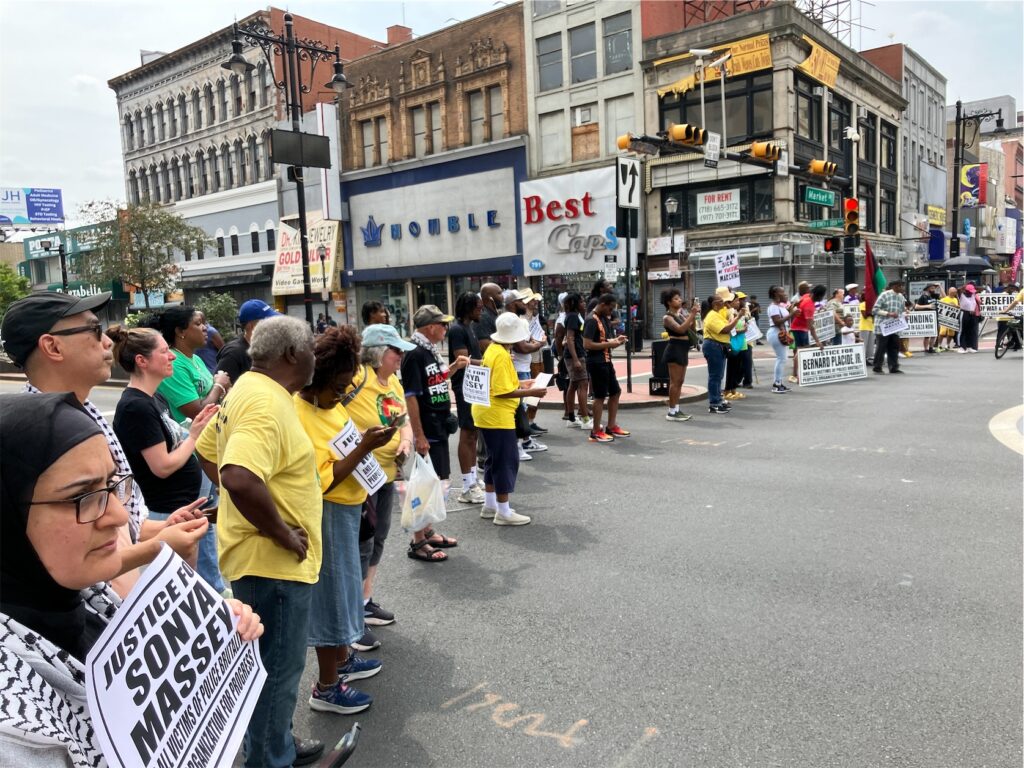 semicircle of activists, many holding signs surrounding intersection of Broad and Market streets in Newark, N.J.