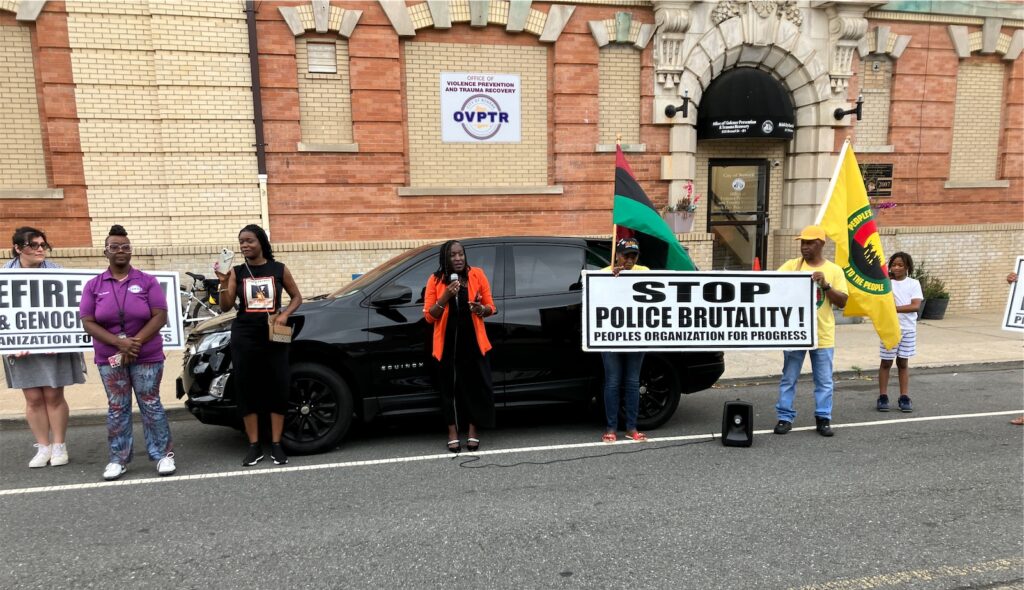 Activists outside Newark’s Office of Violence Prevention and Trauma Recovery at the end of the march; some hold People’s Organization signs, the central one saying “STOP POLICE BRUTALITY!”. The speaker, in black slacks and top with a red jacket, holds a microphone; to her left, along with the sign, activist hold a PanAfrican red-black-green flag and a People’s Organization for Progress flag, with their logo on a yellow field