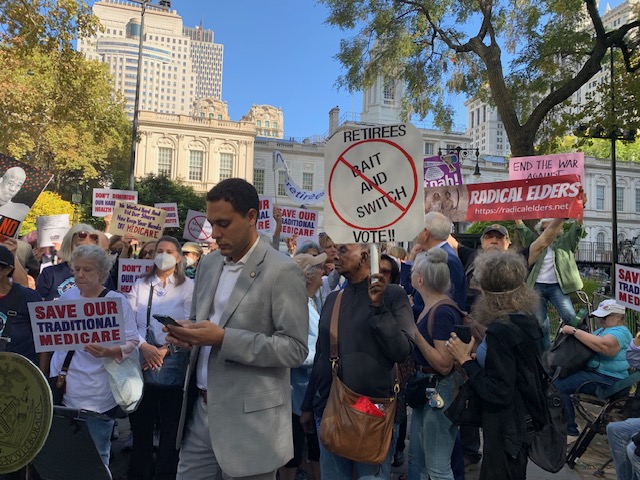 Retired city employees and allies rally outside NYC city hall. Signs read “Save our Traditional Medicare”, “Retirees Vote, No Bait and Switch”, etc.