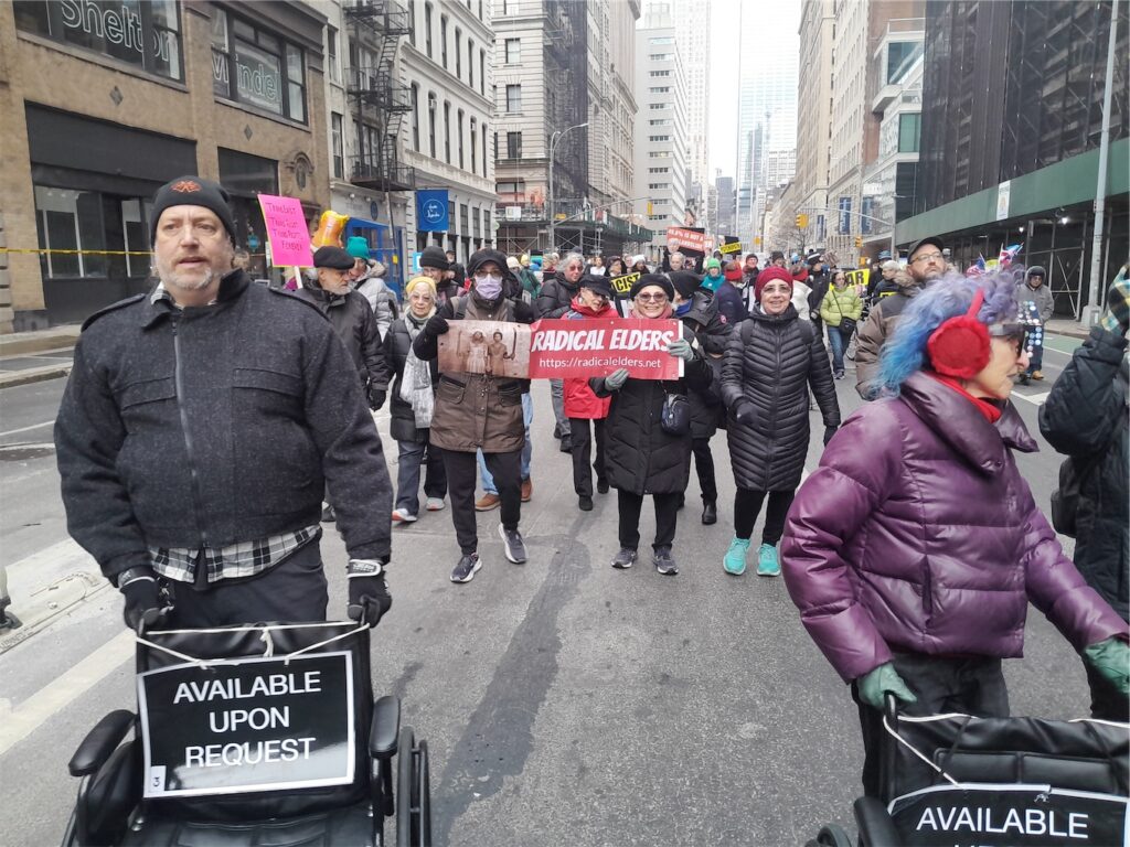 Radical Elders arch with signs, led by 2 people pushing wheelchairs with signs saying “Available Upon Request”.