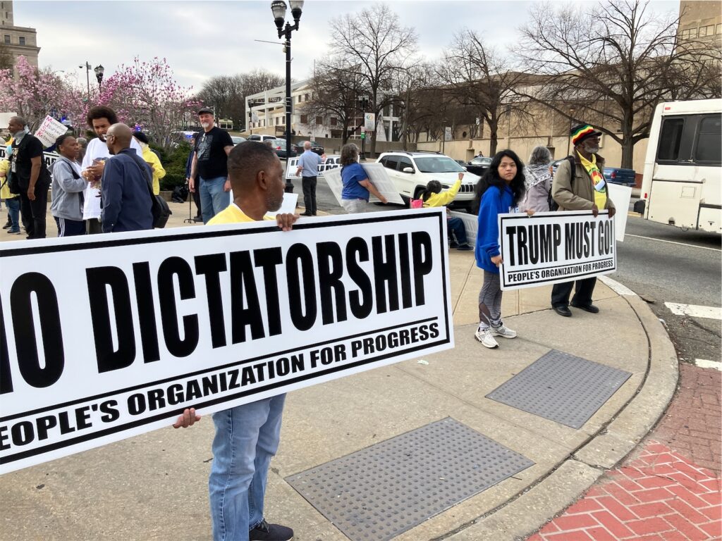 Activists holding People’s Organization for Progress banners on street corner as cars drive by. The messages are “NO DICTATORSHIP” and “TRUMP MUST GO!” More activists in conversation behind those holding banners.