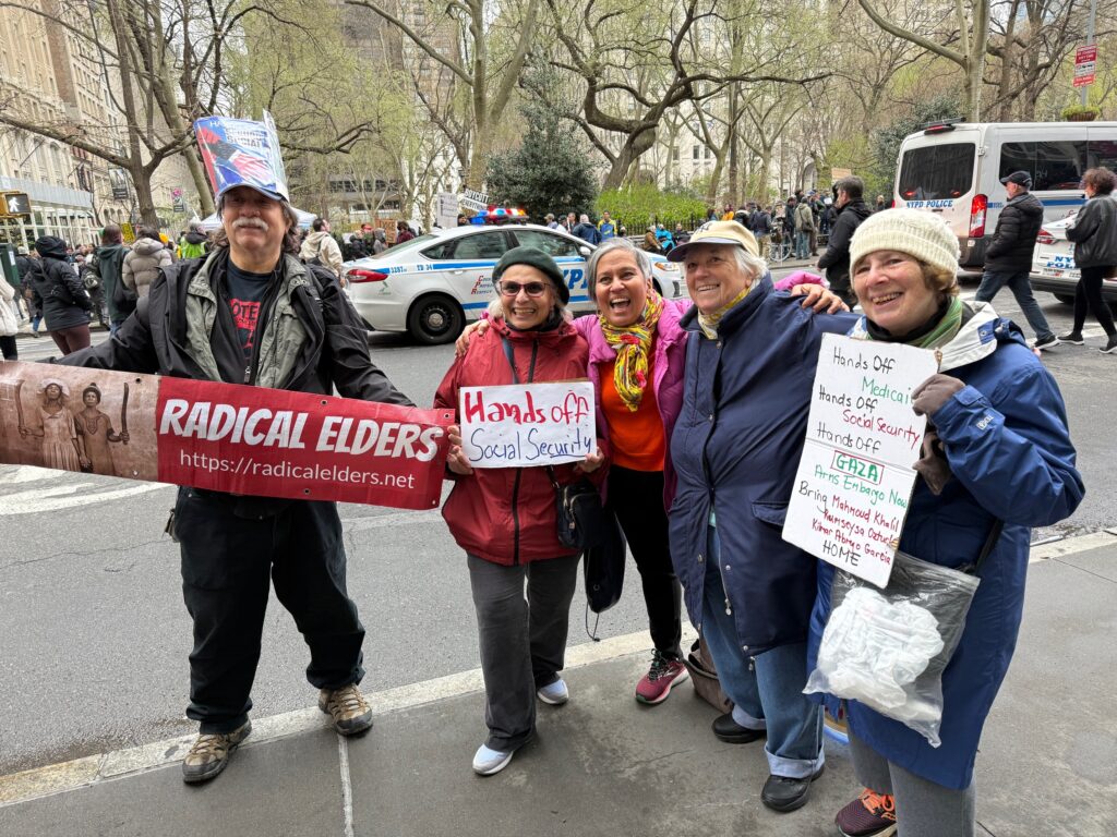Smiling Radical Elders members hold “Hands Off” signs and a Radical Elders banner across the street from police vehicles and the protest crowd in Bryant Park