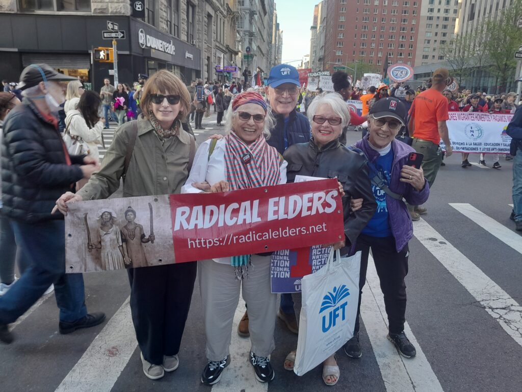 Smiling Radical Elders hold our banner on New York street with marchers holding signs in the background.