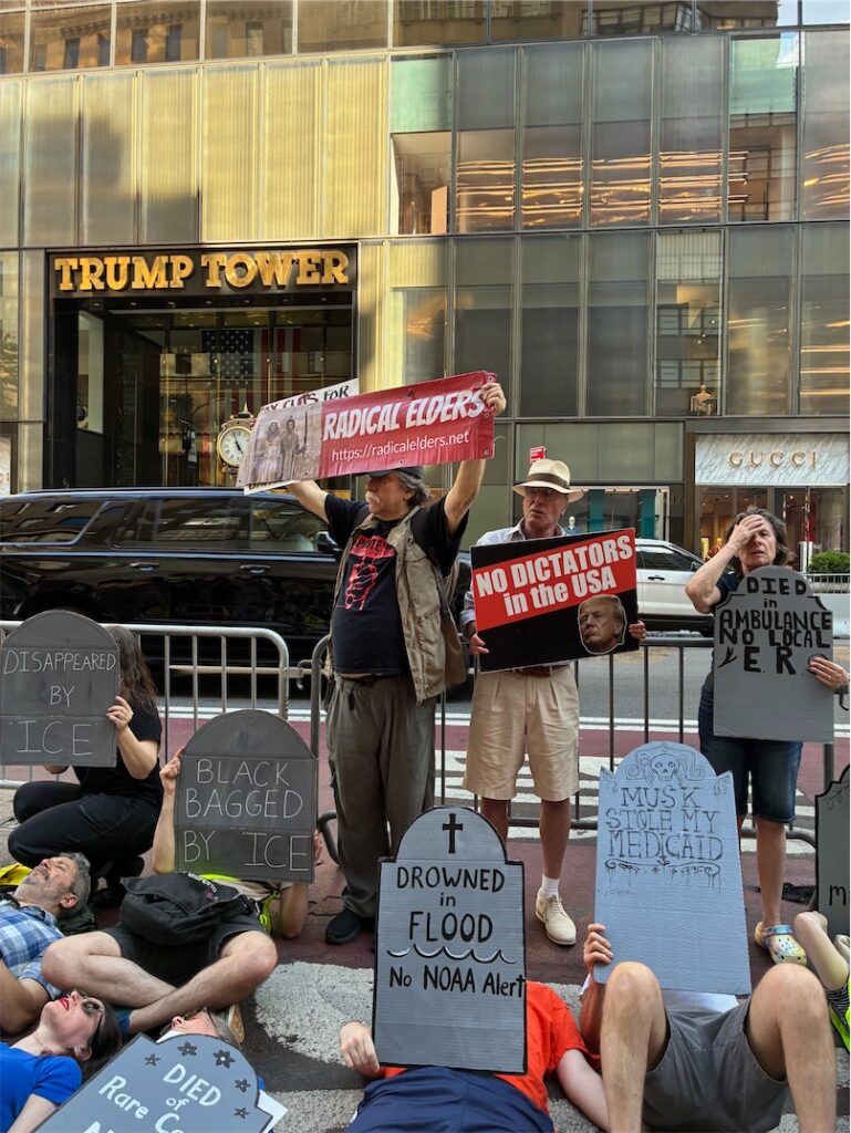 Die-in in front of Trump Tower, most signs shaped like gravestones, also people holding Radical Elders banner and sign saying “No dictators in the USA”.