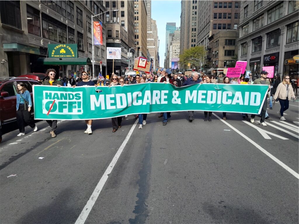 Crowd carrying signs behind a banner reading “Hands Off! Medicare and Medicaid”.