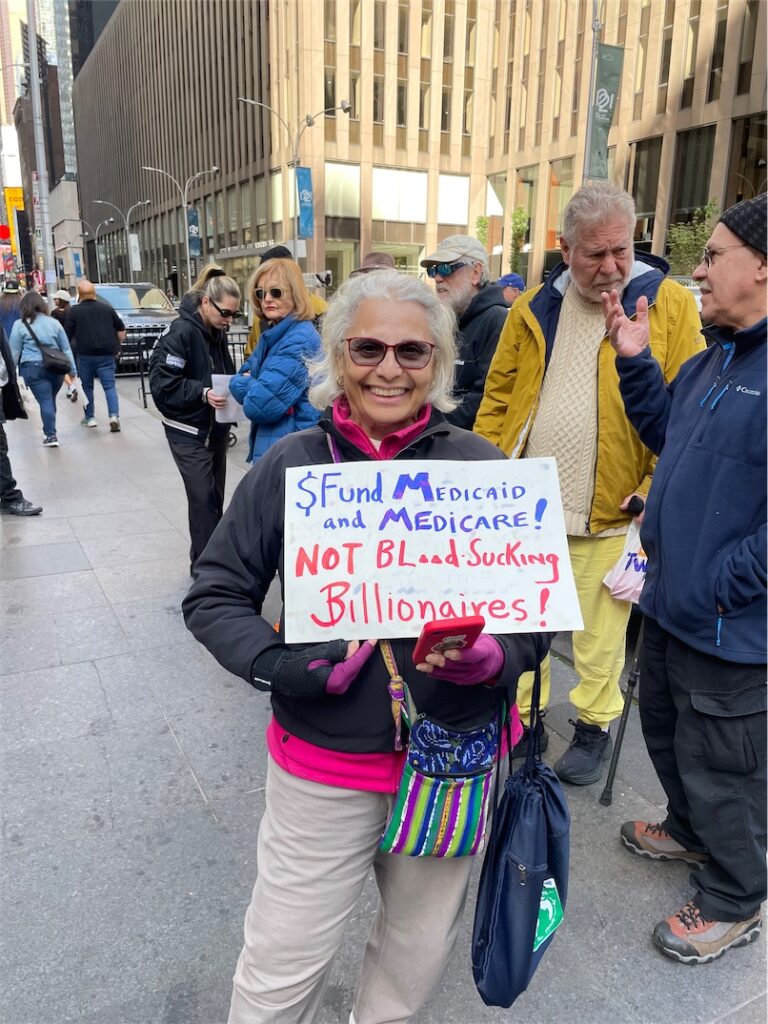 Smiling Radical Elder poses with a homemade sign reading $ Fund Medicaid and Medicare! Not Bloodsucking Billionaires!