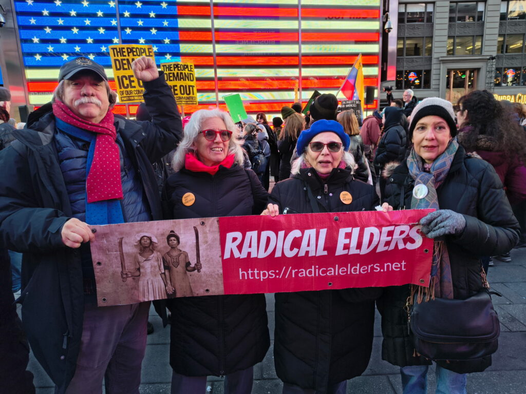 Radical Elders hold an RE banner in front of a giant wooden USA flag . Other ralliers hold signs saying The People Demand ICE Out of Our Communities, a Venezuelan flag, and more