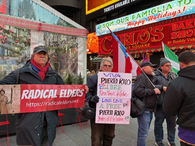 Radical Elders hold an RE banner and sign reading Viva Puerto Rico Libre in Spanish and English; others hold flags of other countries; all in front of Carlo’s Bake Shop in Times Square