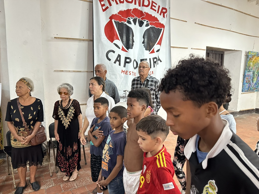 Elders and young boys gathered in front of a poster (illegible—two faces creating the silhouette of a tree between them) on what appears to be a gallery wall.