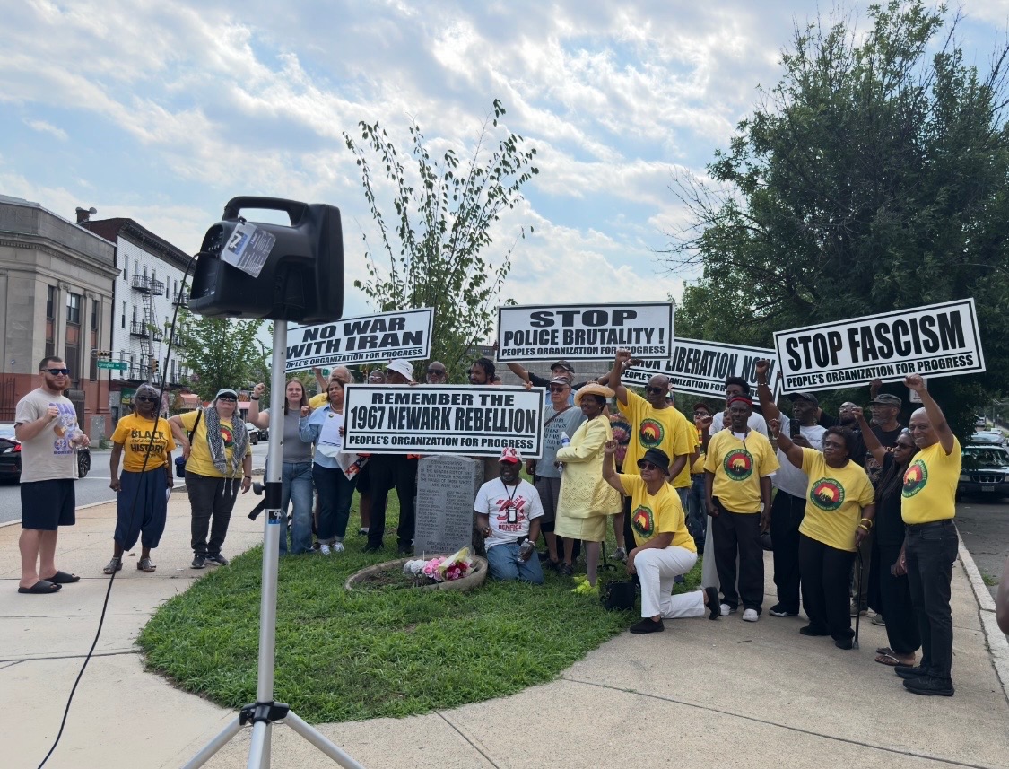 Group of people, most wearing People’s Organization for Progress (POP) yellow T-shirts and many holding POP signs, with the central sign reading “Remember the 1967 Newark Rebellion / People’s Organization for Progress”, gathered in front of an ABC news camera on a sidewalk and grass median on a summer day