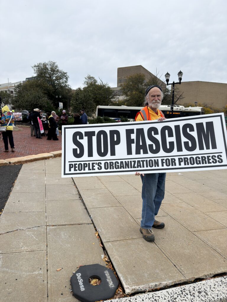 Kevin Norton holds a People’s Organization for Progress sign reading Stop fascism. Other activists gather in the background.