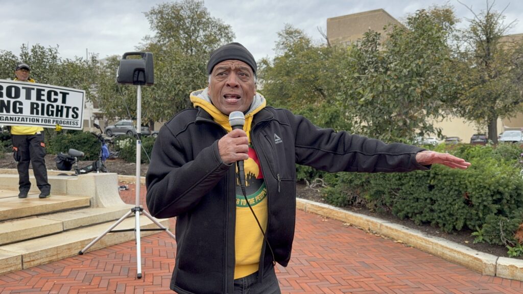 Larry Hamm speaks into a microphone at the No Kings event in Newark. Behind him someone holds a sign reading Protect Voting Rights, People’s Organization for Progress.