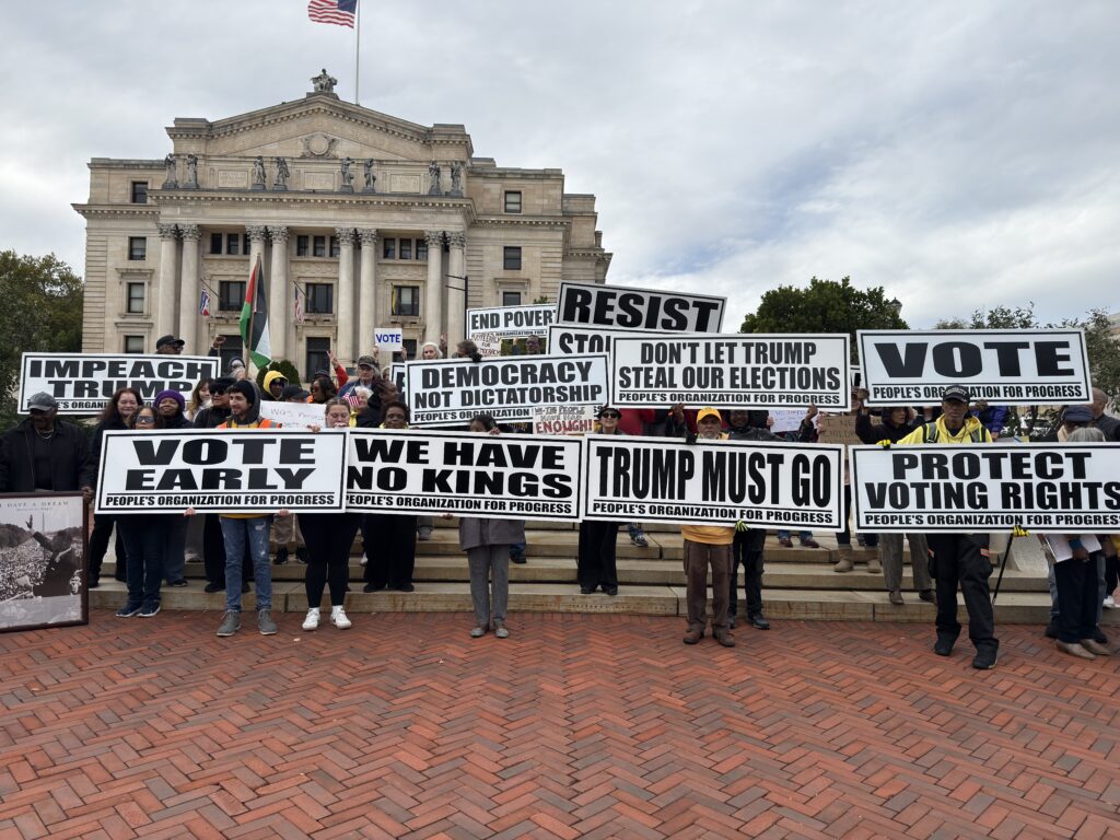 Activists hold 11 People’s Organization for Progress signs in front of the Essex County Courthouse. The signs read (first row): Vote Early, We Have No Kings, Trump Must Go, Protect Voting Rights, (second row): Impeach Trump, Democracy Not Dictatorship, Don’t Let Trump Steal Our Elections, VOTE. Behind these are Resist and End Poverty.