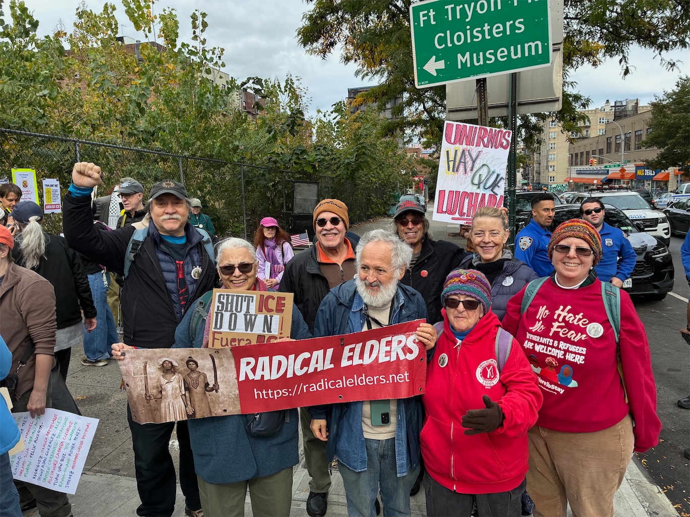 Smiling Radical Elders in warm clothes pose near a Fort Tryon Park/Cloisters Museum sign holding a RE banner and signs reading Shut Down ICE and Unirnos Hay Que Luchar. Two police officers are visible behind them.